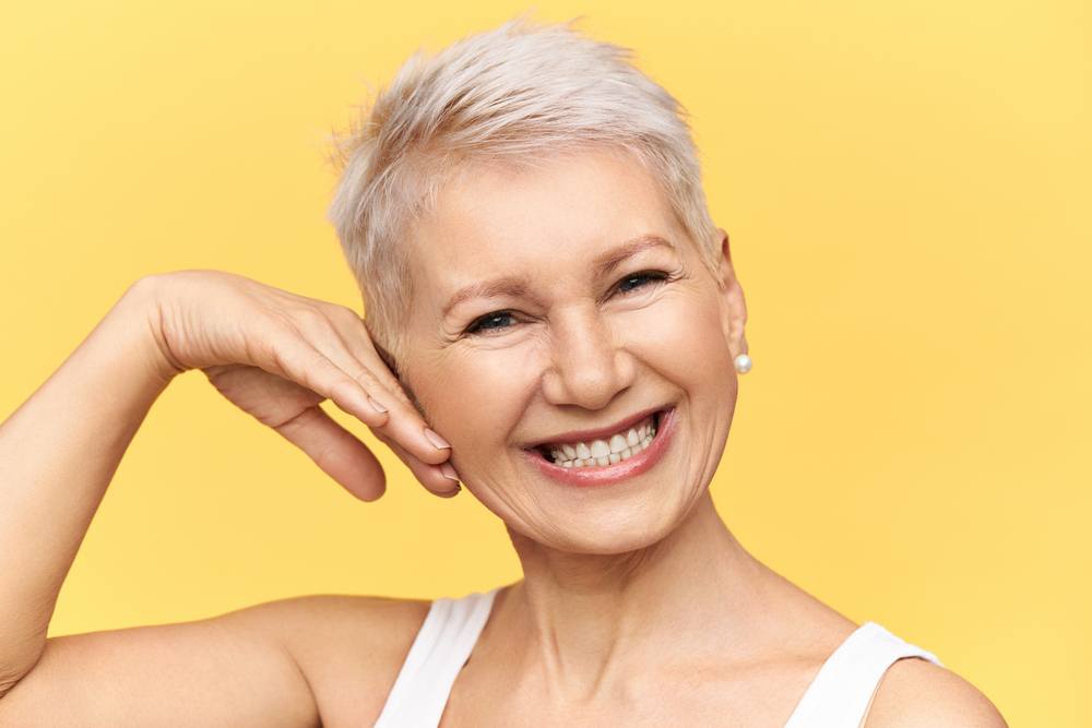 Studio shot of charismatic positive middle aged female posing against yellow background touching cheek, looking at camera with broad cheerful smile, taking care of her wrinkled skin, applying cream Протезування зубів в Одесі