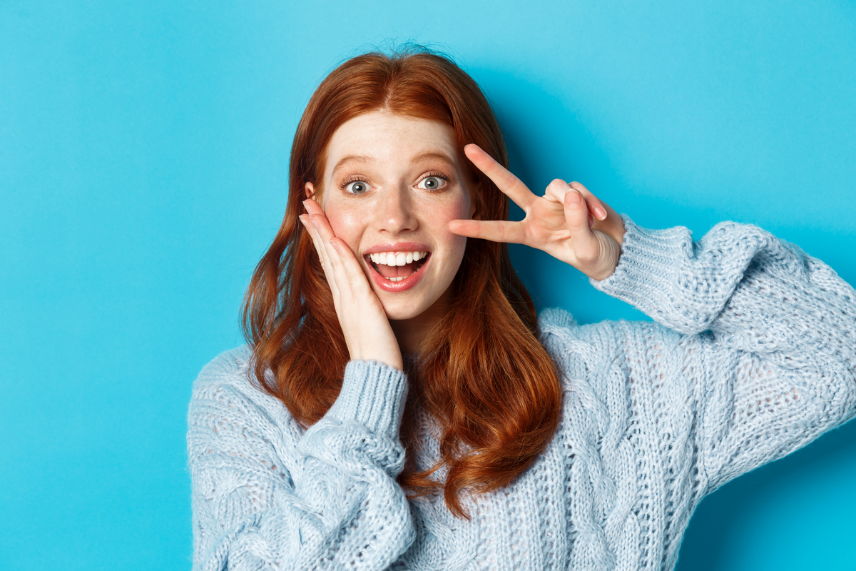 Close-up of beautiful smiling girl with red hair, showing peace kawaii sign and gazing at camera, standing over blue background Домашнє відбілювання зубів